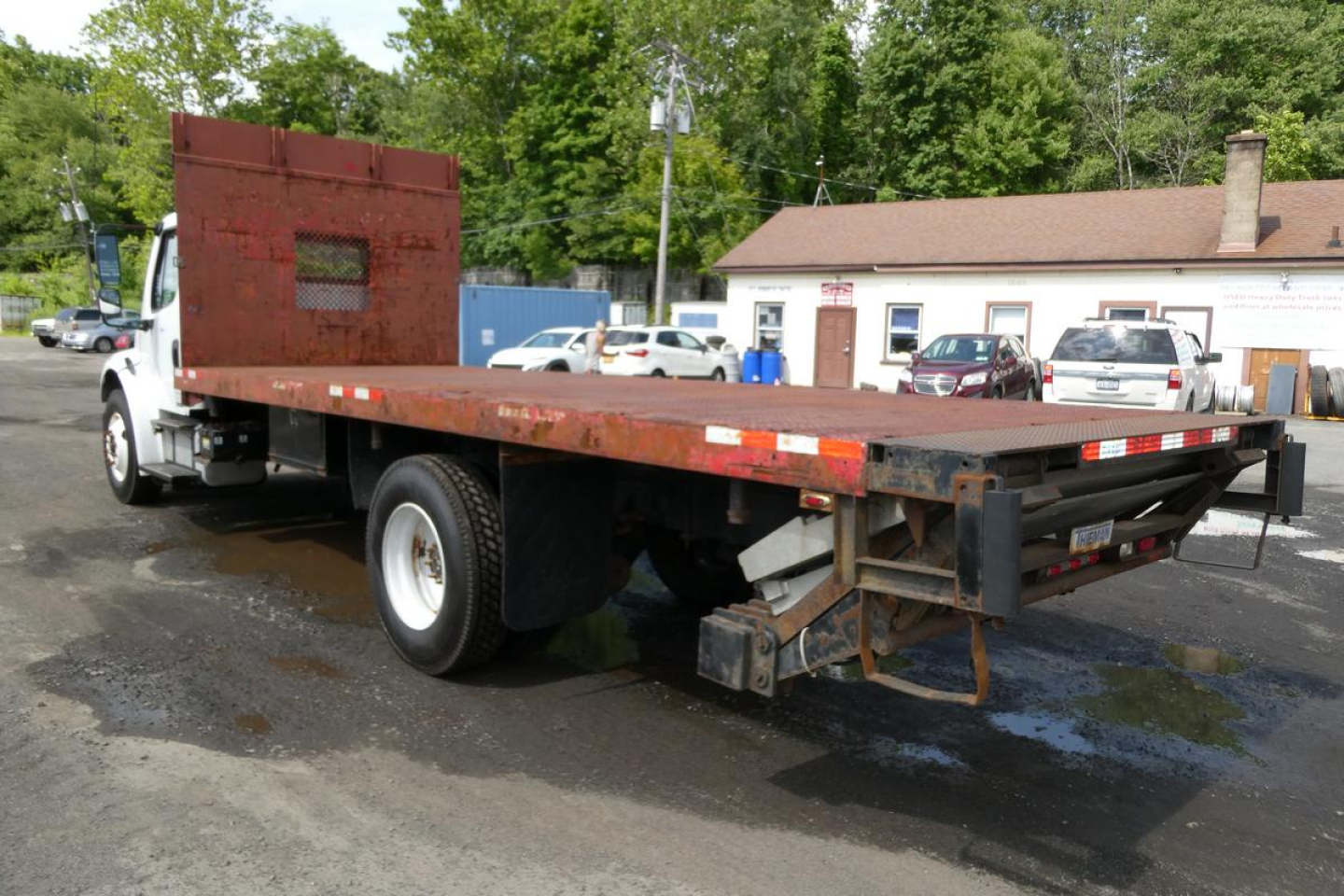 Flatbed truck with wooden deck and red metal cab shield showing a Freightliner M2-106 flatbed truck. Side panels reflectors and a rear deck with mechanical components visible.