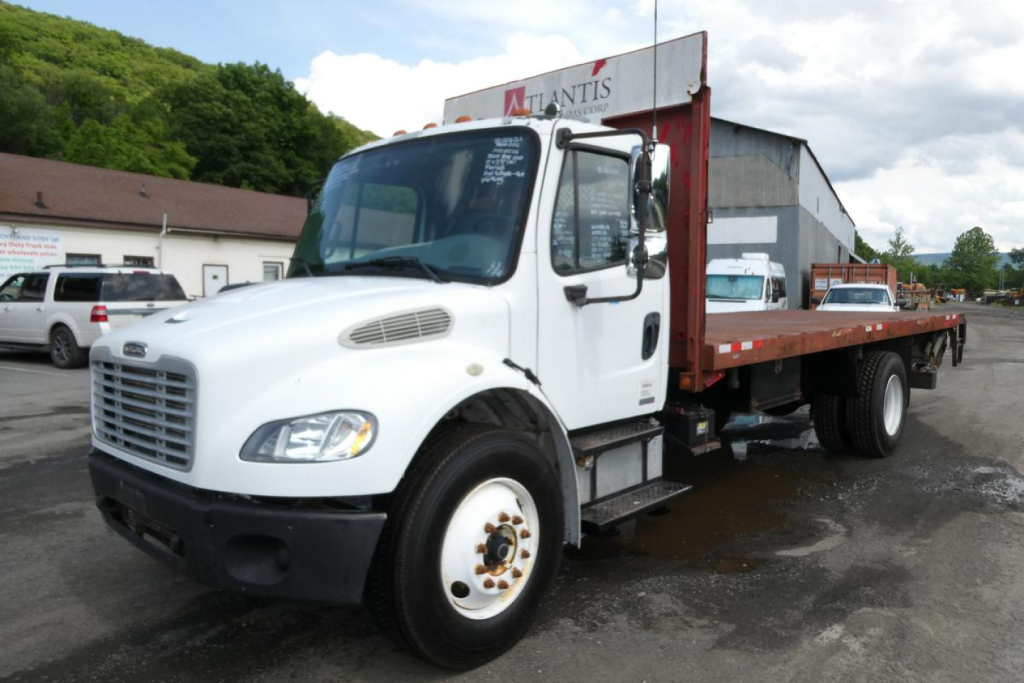 Flatbed tow truck showing Freightliner M2-106 cab and long cargo bed with ramp in a dealership lot.