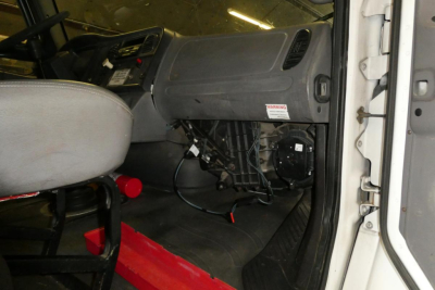 Freightliner M2-106 cab interior showing dashboard, driver seat, and exposed wiring under dash with warning label on glove box.