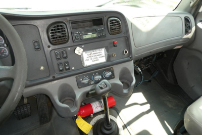 Freightliner M2-106 dashboard and center console showing steering wheel, radio, climate controls, and a red fire extinguisher under the gear shifter.