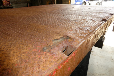 Flatbed truck bed with rusty, diamond-plate surface and damaged edge, visible corrosion and puncture near corner.