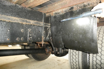 Freightliner M2-106 chassis view showing undercarriage, frame rails, rear axle, leaf springs, and mud flap with a hand holding a tool near the wheel.