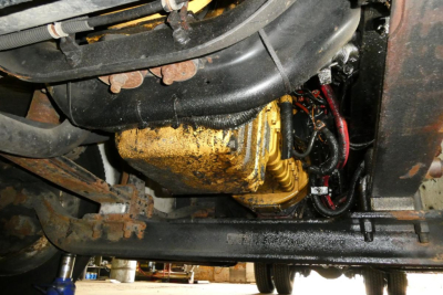 Underbelly view of a Freightliner M2-106 engine area showing a gold-colored component with hoses and wiring, framed by black metal chassis.