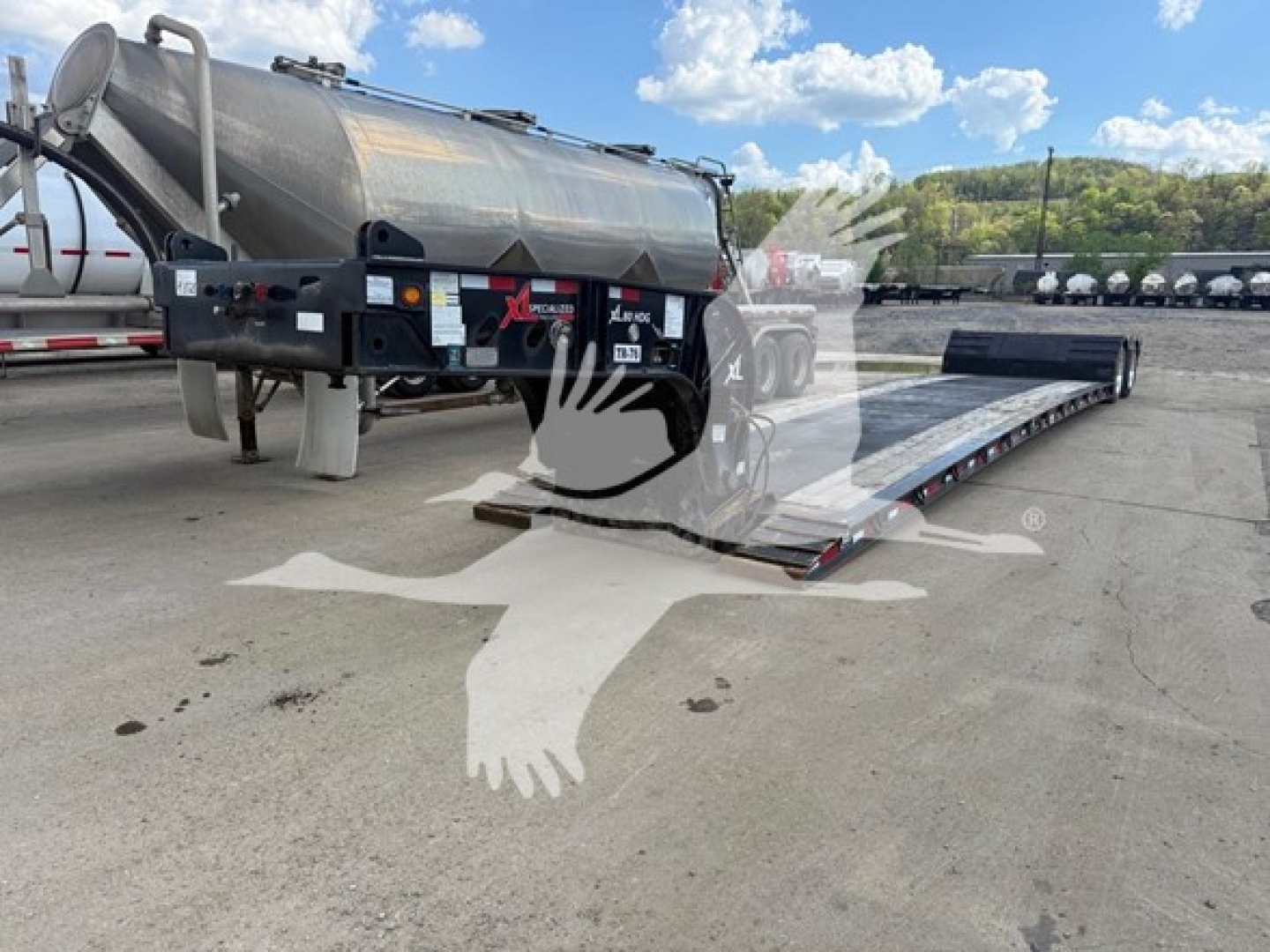 Trailer with extendable lowboy ramp in a loading yard, side view showing cylindrical tank and dual-axle trailer with hydraulic platform. Visible labels include stock number and VIN area.