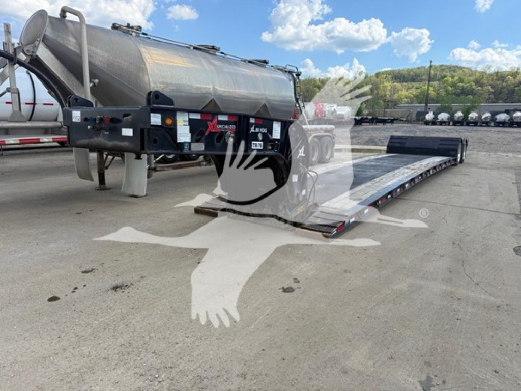 Trailer with extendable lowboy ramp in a loading yard, side view showing cylindrical tank and dual-axle trailer with hydraulic platform. Visible labels include stock number and VIN area.