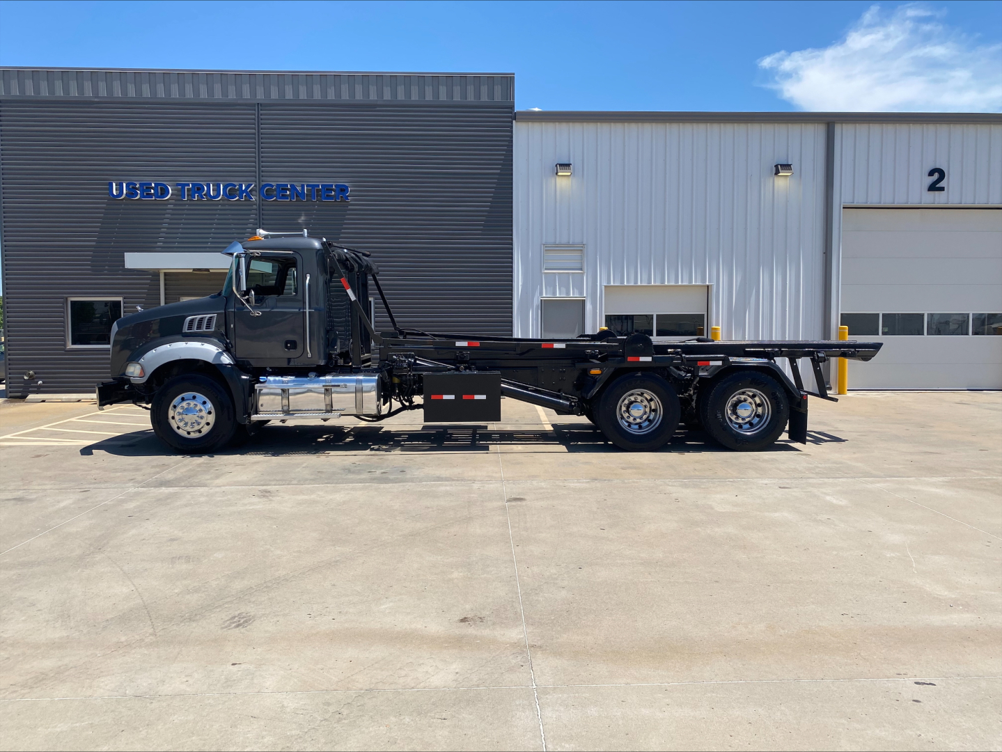 Side view of a 2016 Mack GU533 roll-off truck showing cab, chassis, and long flatbed with dual rear axles parked in front of a building labeled USED TRUCK CENTER.