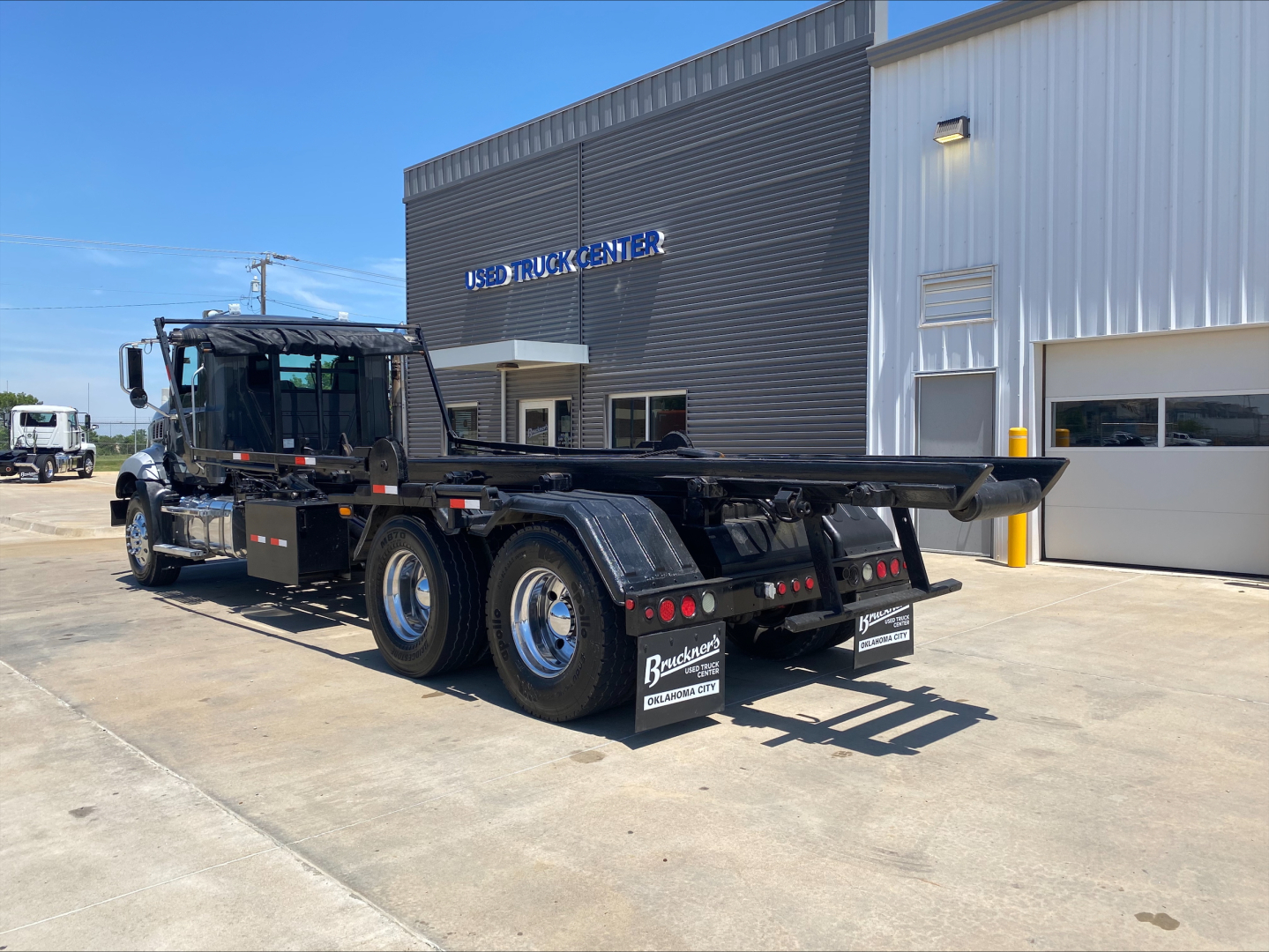 Truck rear and cab of a 2016 Mack GU533 roll-off truck showing dual rear axles and flatbed with black mud flaps and Forks. Visible text includes dealership signage on building.