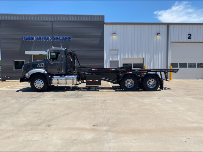 Side view of a 2016 Mack GU533 roll-off truck showing cab, chassis, and long flatbed with dual rear axles parked in front of a building labeled USED TRUCK CENTER.