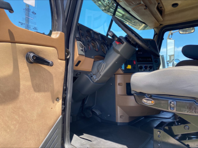 Truck cab interior of a 2016 Mack GU533 rolloff showing steering wheel, dashboard gauges, beige door panel, seat with tag, and pedals.