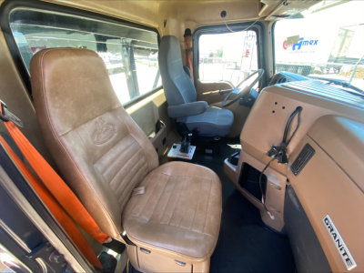 Truck cab interior showing steering wheel, driver seat, and dashboard of a Mack GU533 roll-off truck; beige/brown seats, gray floor mat, stock number badge visible.