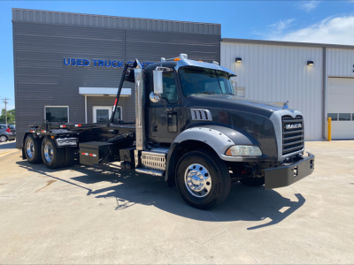 Mack GU533 roll-off truck in front of dealership showing cab and flatbed with hydraulic arms and wheel setup.
