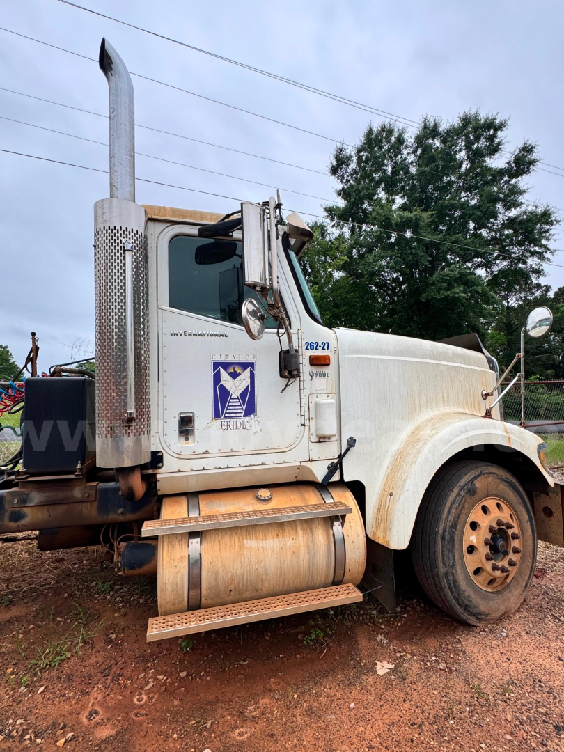 Truck side view of a white 2007 International 9900i rolloff truck showing exhaust stack, front fender, and driver's door with logo and stock number plate.
