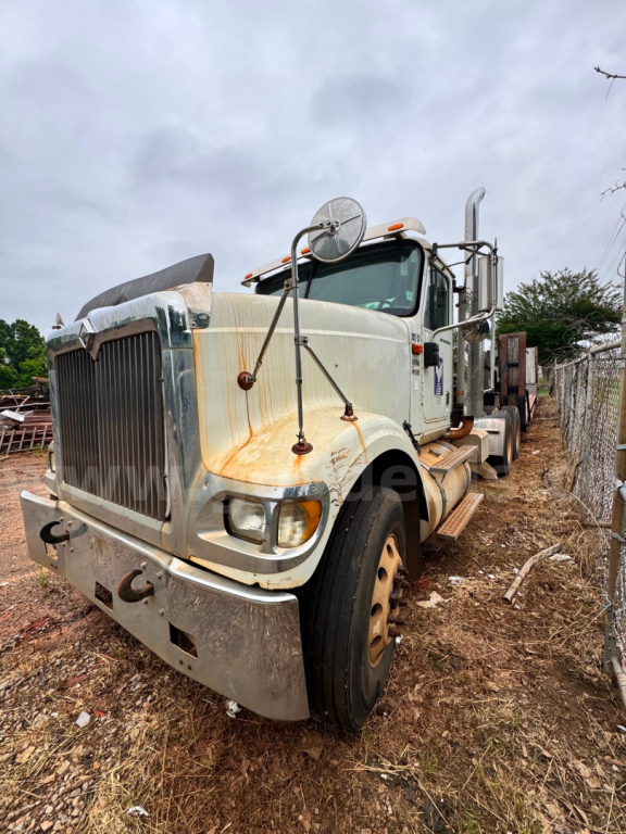 Heavy-duty truck cab view of a white 2007 International 9900i with rust accents, large grille, side mirrors, and sleeper cab; front wheels on dirt at yard.