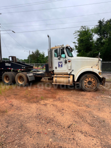 Truck side-view of a white 2007 International 9900i roll-off truck with a flatbed trailer and brownish dirt ground.