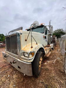 Heavy-duty truck cab view of a white 2007 International 9900i with rust accents, large grille, side mirrors, and sleeper cab; front wheels on dirt at yard.