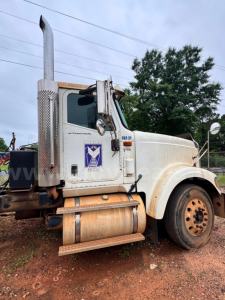 Truck side view of a white 2007 International 9900i rolloff truck showing exhaust stack, front fender, and driver's door with logo and stock number plate.