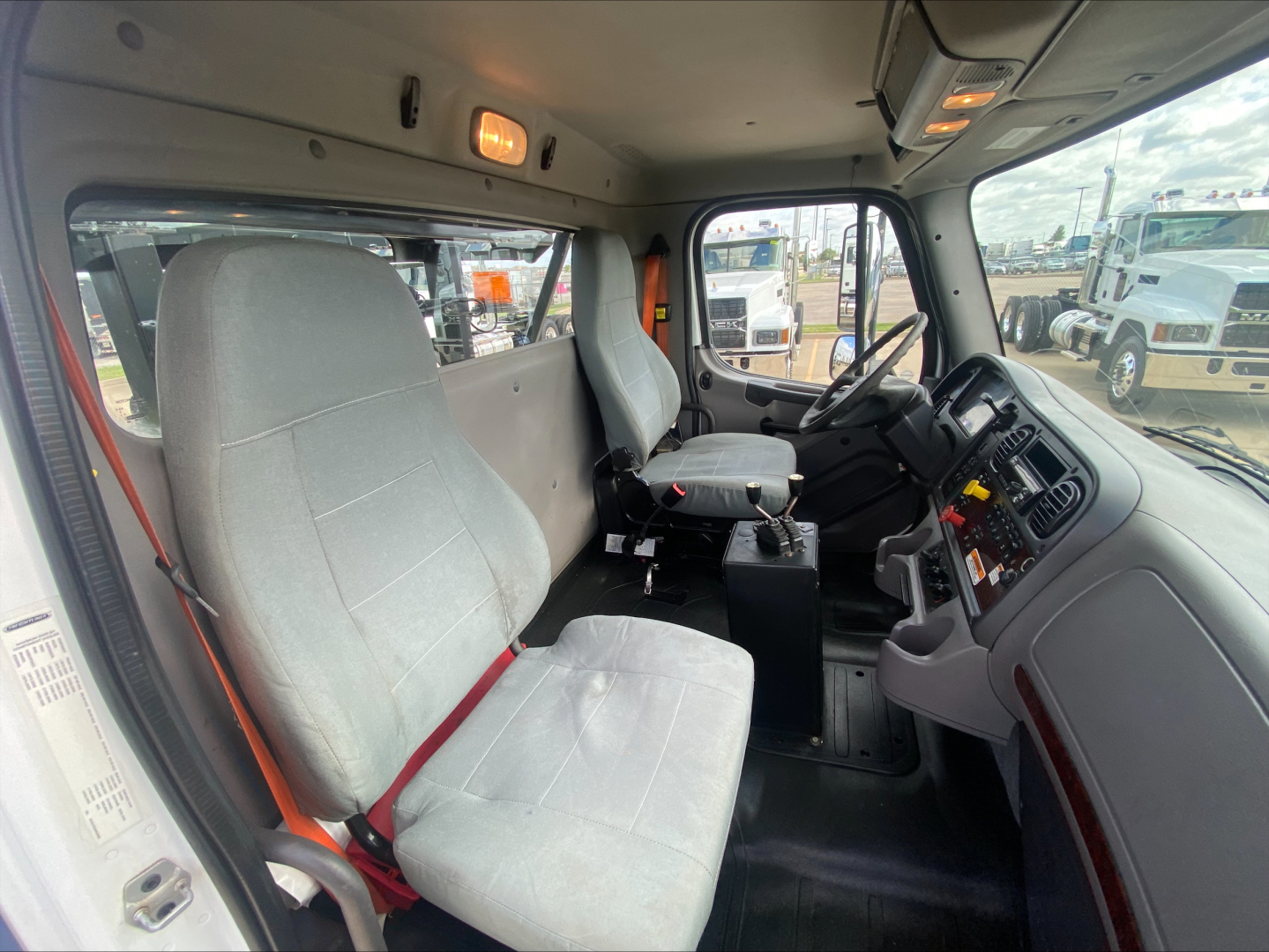 Freightliner M2 2019 cab interior with two gray fabric seats, steering wheel and dashboard visible; driver seat has red seat belt.