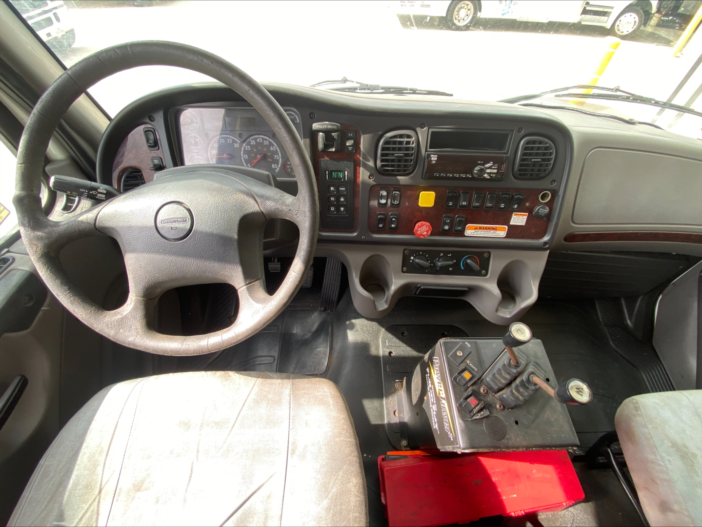 Dashboard and steering wheel of a Freightliner M2 cab showing steering wheel, instrument cluster, center stack, and gear shifter; red hydraulic control box on floor.