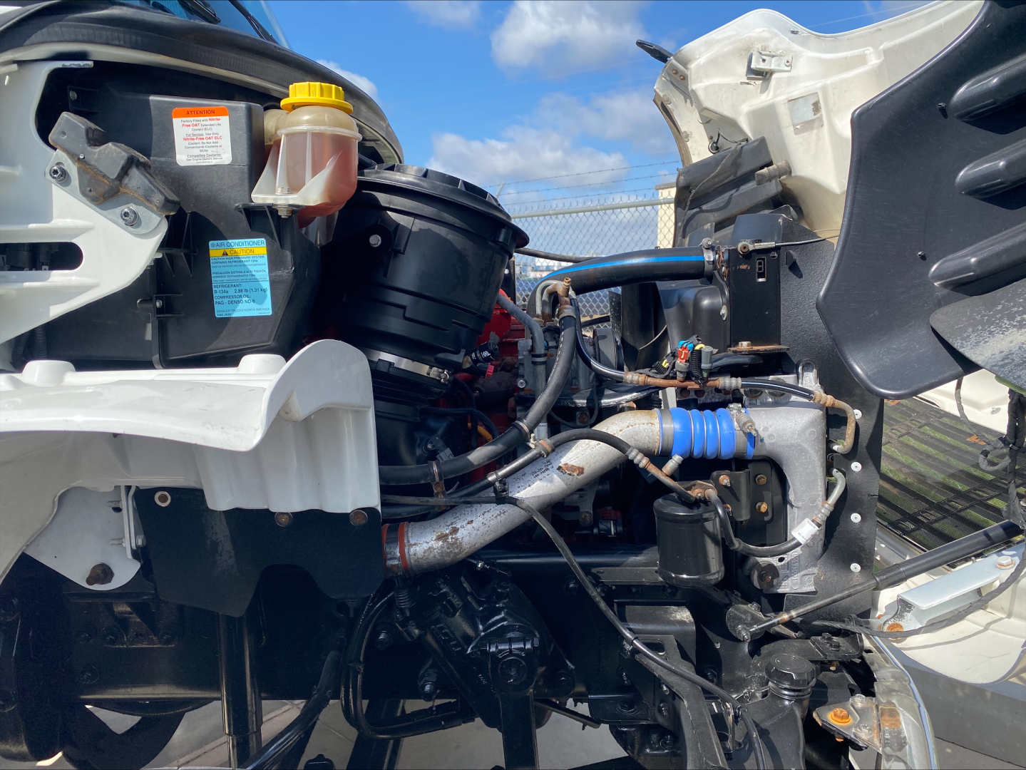 Engine bay of a Freightliner M2 roll-off truck showing hydraulic fluid reservoir caps, exposed hoses, and engine components.
