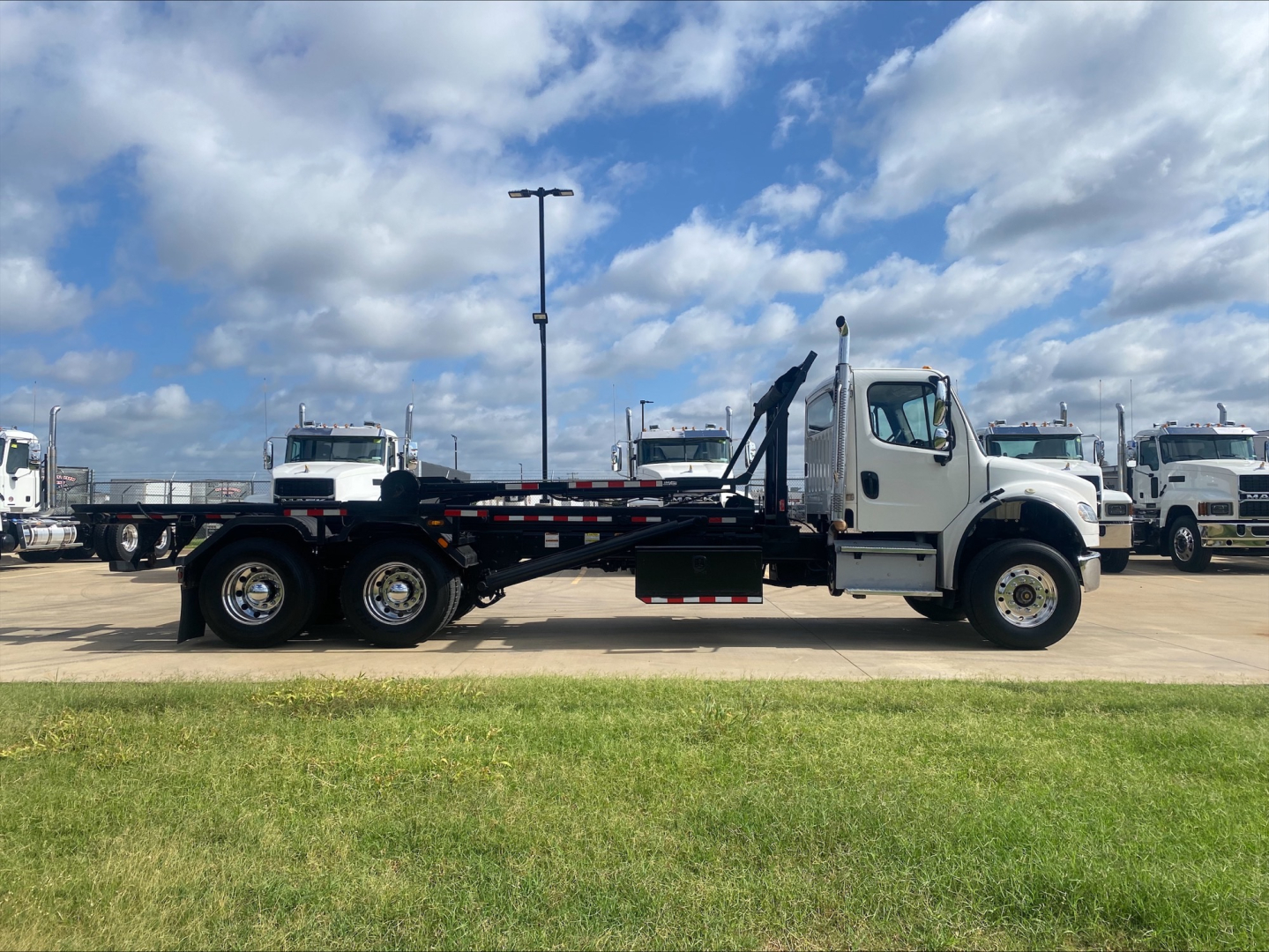 2019 Freightliner M2 rolloff truck side view showing cab, chassis, dual rear axles, and flatbed with hoist in a dealership lot.
