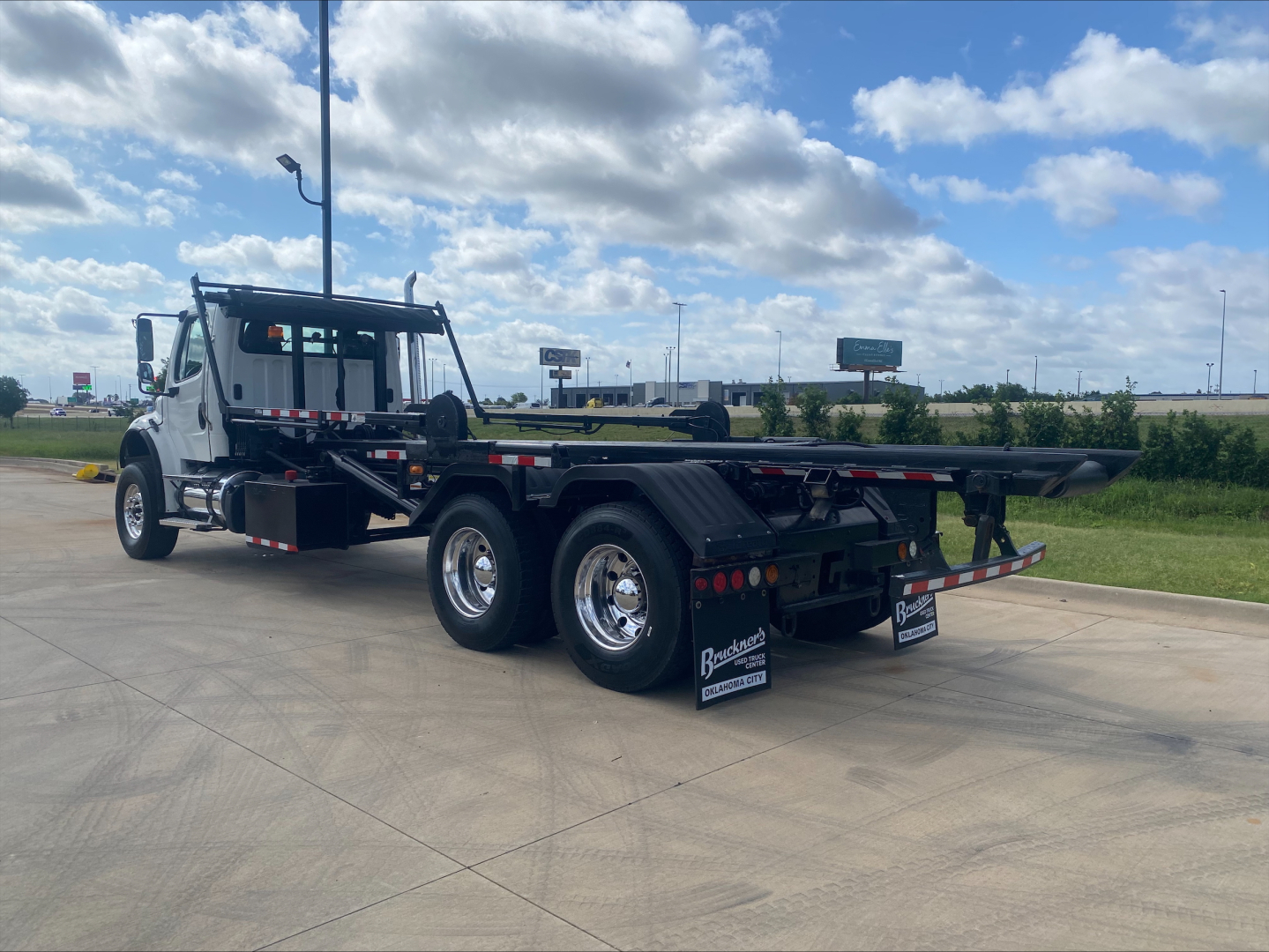 Front-left side of a white 2019 Freightliner M2 roll-off truck with a flatbed dumping bed, visible wheels, and rear decals. Text on mudflaps and side panels shows dealership branding.