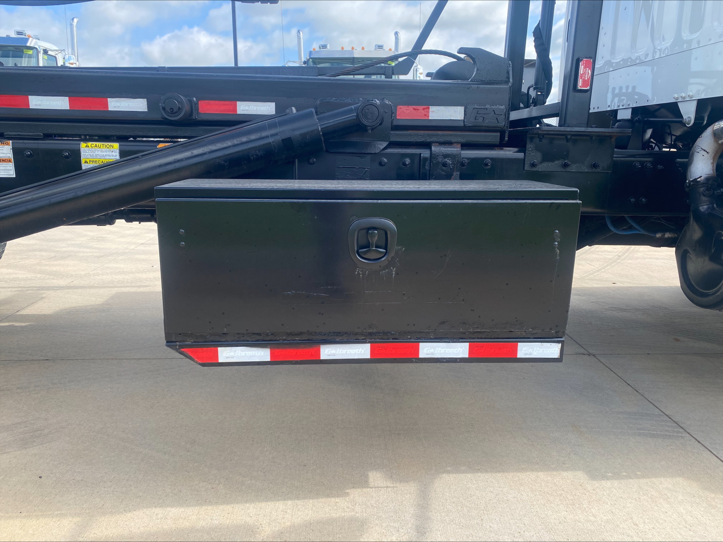 Truck undercarriage showing a black toolbox mounted beneath a Freightliner M2 roll-off body with red-white reflective tape.