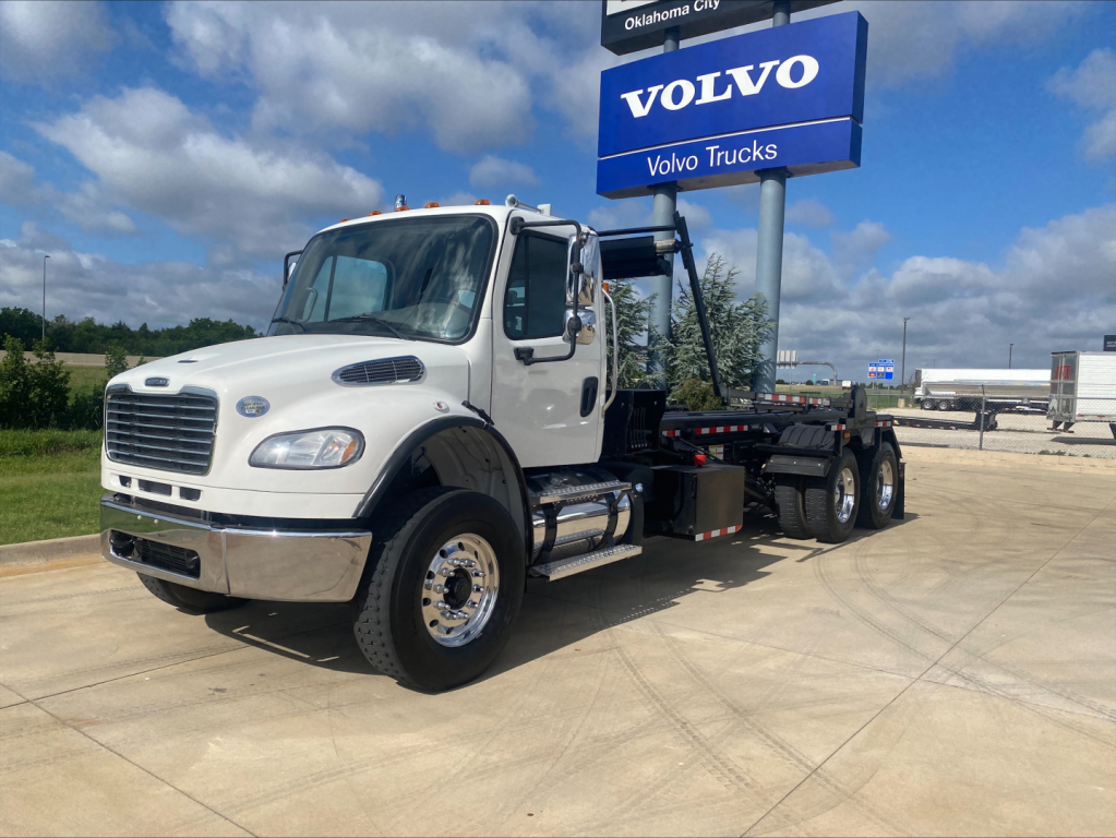2019 Freightliner M2 roll-off truck in gravel lot with Volvo sign in background, white cab, black chassis, several wheels