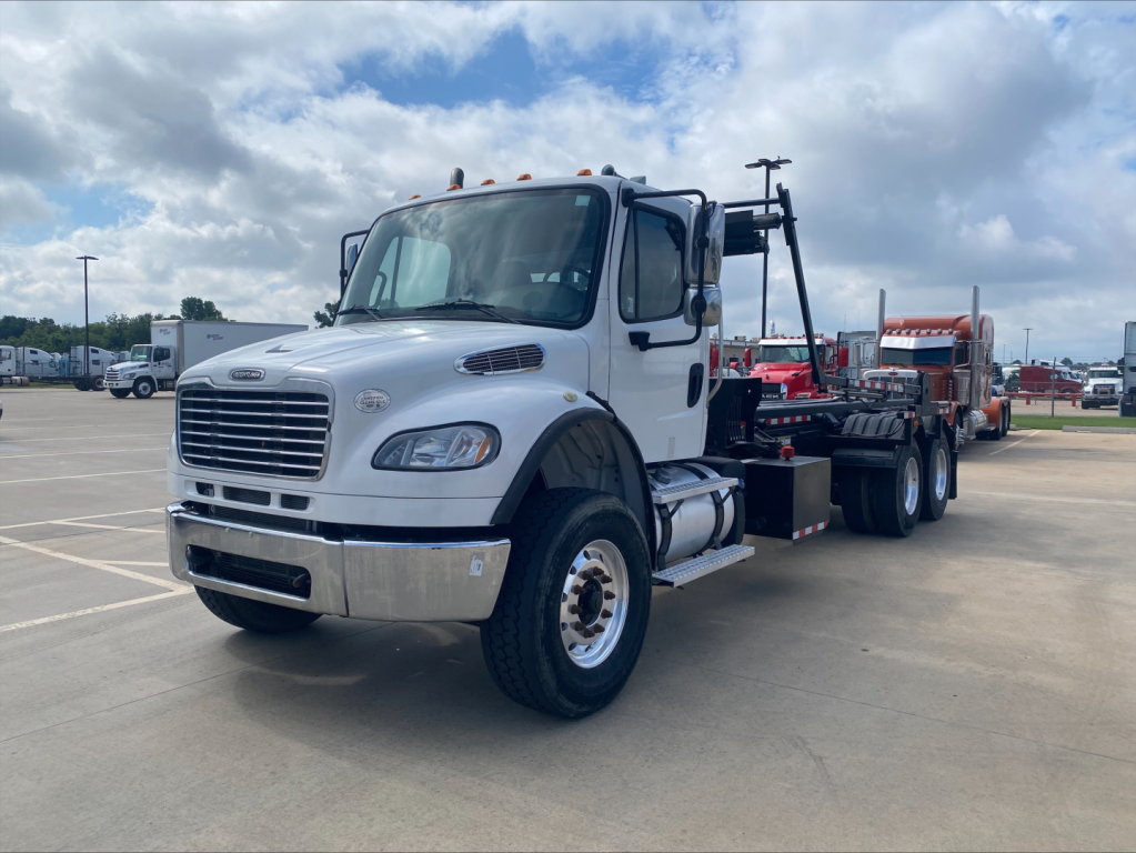 Freightliner M2 rolloff truck in a parking lot, front-left view showing cab, white exterior, chrome bumper, and attached trailer chassis.