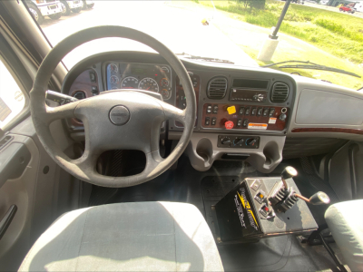 Dashboard of a Freightliner M2 cab with steering wheel, gauges, and control panel; gear shift/joystick and a secondary control panel are visible.