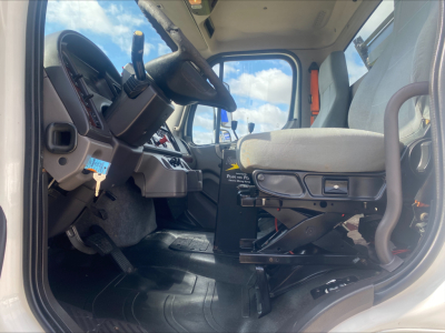 Freightliner M2 truck cab interior showing steering wheel, dashboard, gear selector, and driver's seat with light-gray upholstery.