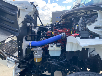 Engine bay of a 2019 Freightliner M2 showing blue hose, white coolant reservoir, black hoses, and red engine components in a roll-off truck configuration.