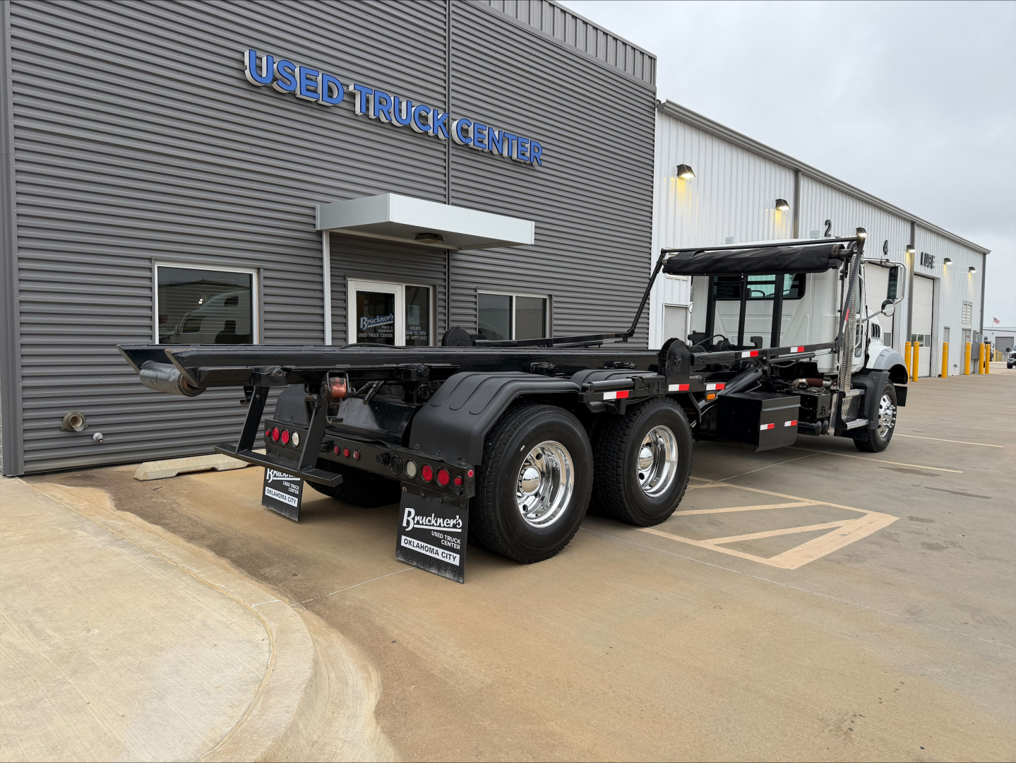 rear view of a Mack GU533 roll-off truck with a flatbed and multiple axles, parked outside a Used Truck Center showroom.