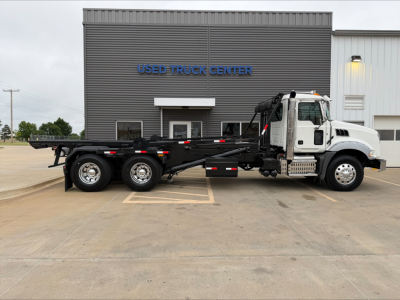 2016 Mack GU533 roll-off truck in front of Used Truck Center building. Side view shows flatbed with three rear axles and a white cab. Building reads 'USED TRUCK CENTER' in blue letters.