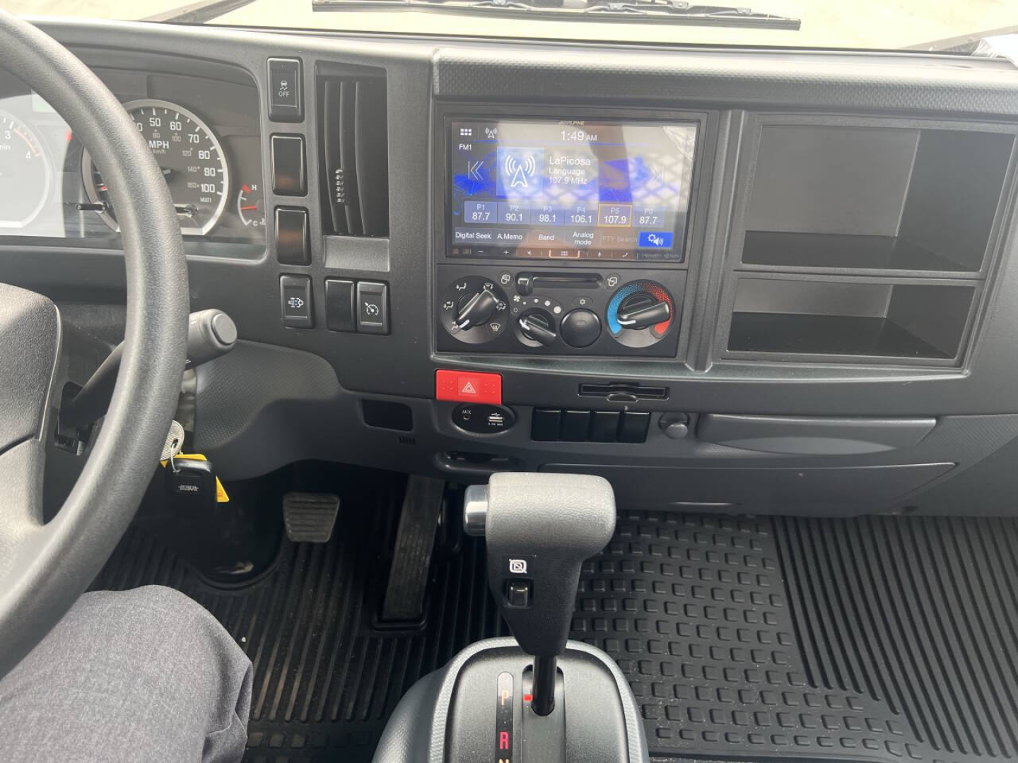 Dashboard view of a 2023 Chevrolet 4500 HD flatbed truck showing steering wheel, digital display, center console with climate controls, gear shifter, and textured floor mat.
