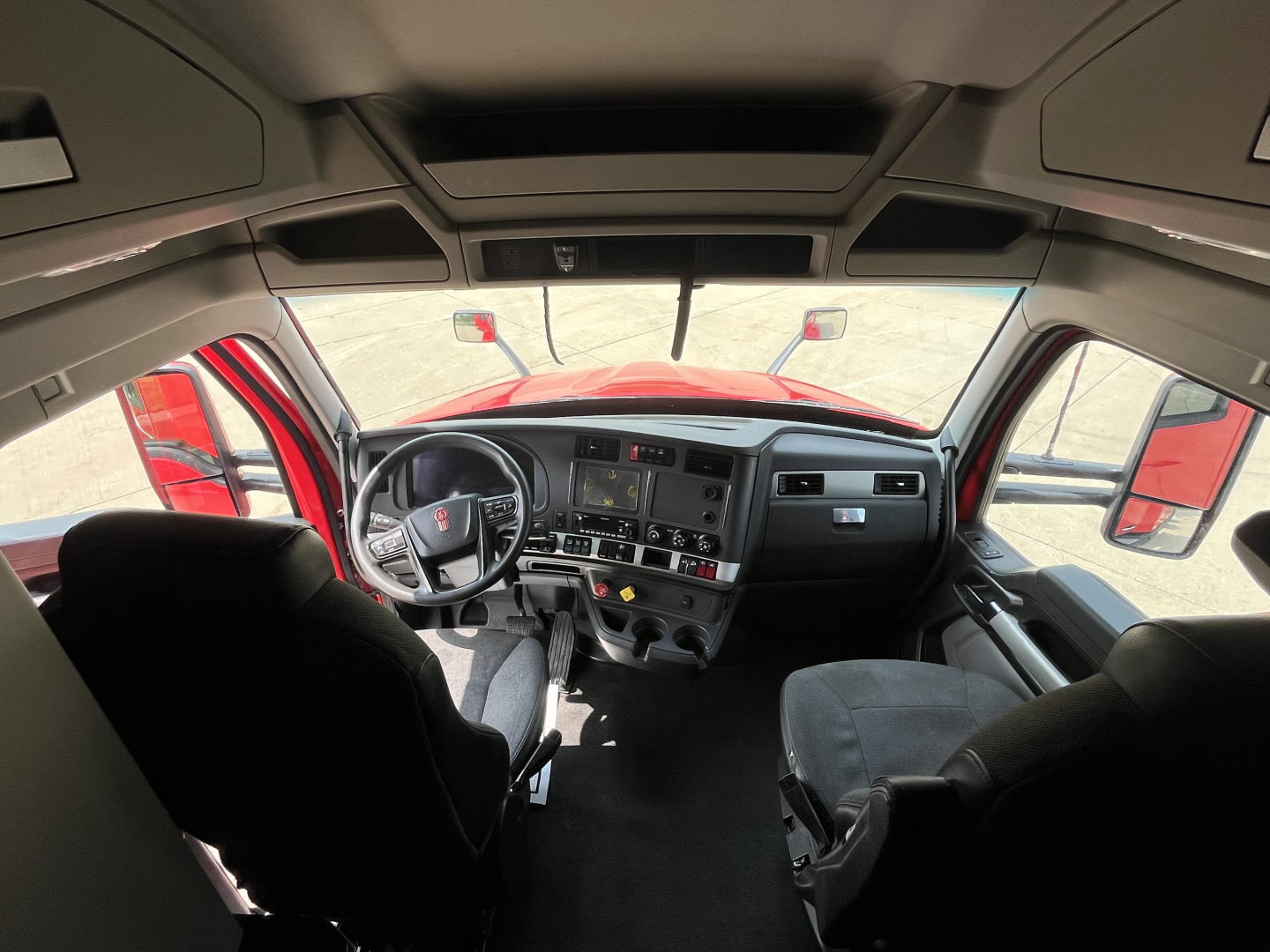 Kenworth T680 cabin interior viewed from behind the seats; dashboard with steering wheel, center console, and overhead storage visible.