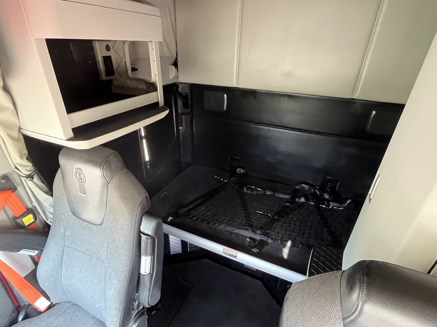 Kenworth T680 cab interior showing sleeper with storage cabinet and padded bed area, driver seat in foreground, black floor matting and seat belt visible.