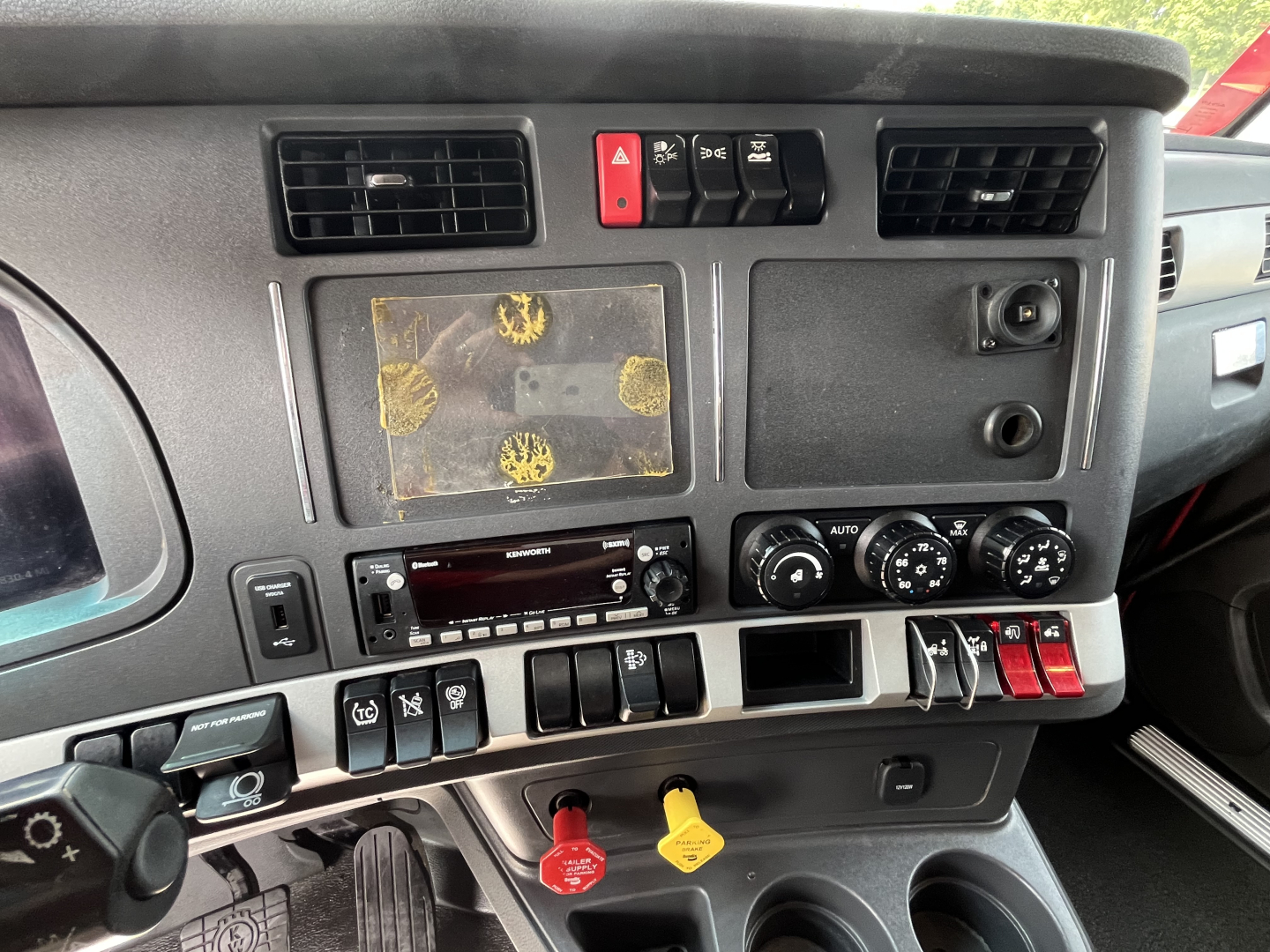 Kenworth T680 dash view showing steering wheel, dashboard vents, control switches, and central display with worn touchscreen area; multiple knobs and red/yellow latches visible.