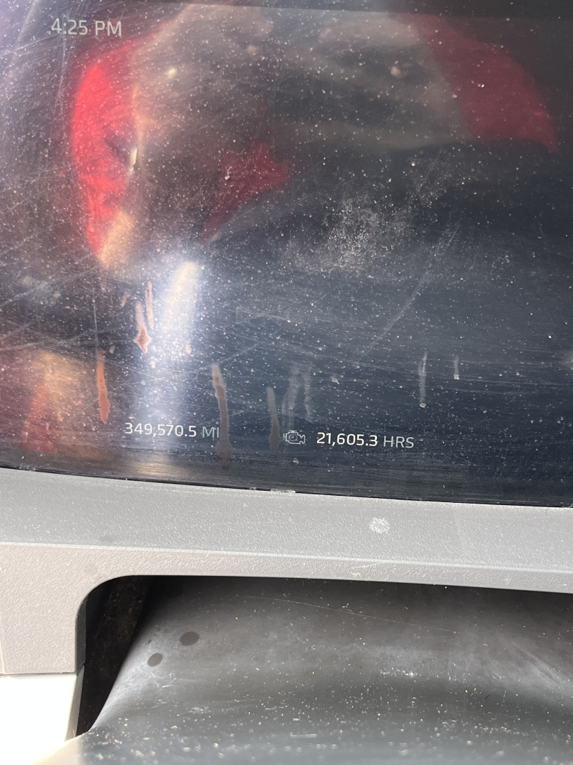 Kenworth T680 dashboard display showing metrics: 4:25 PM, 349,570.5 miles, 21,605.3 hours. Reflection and dust on screen.