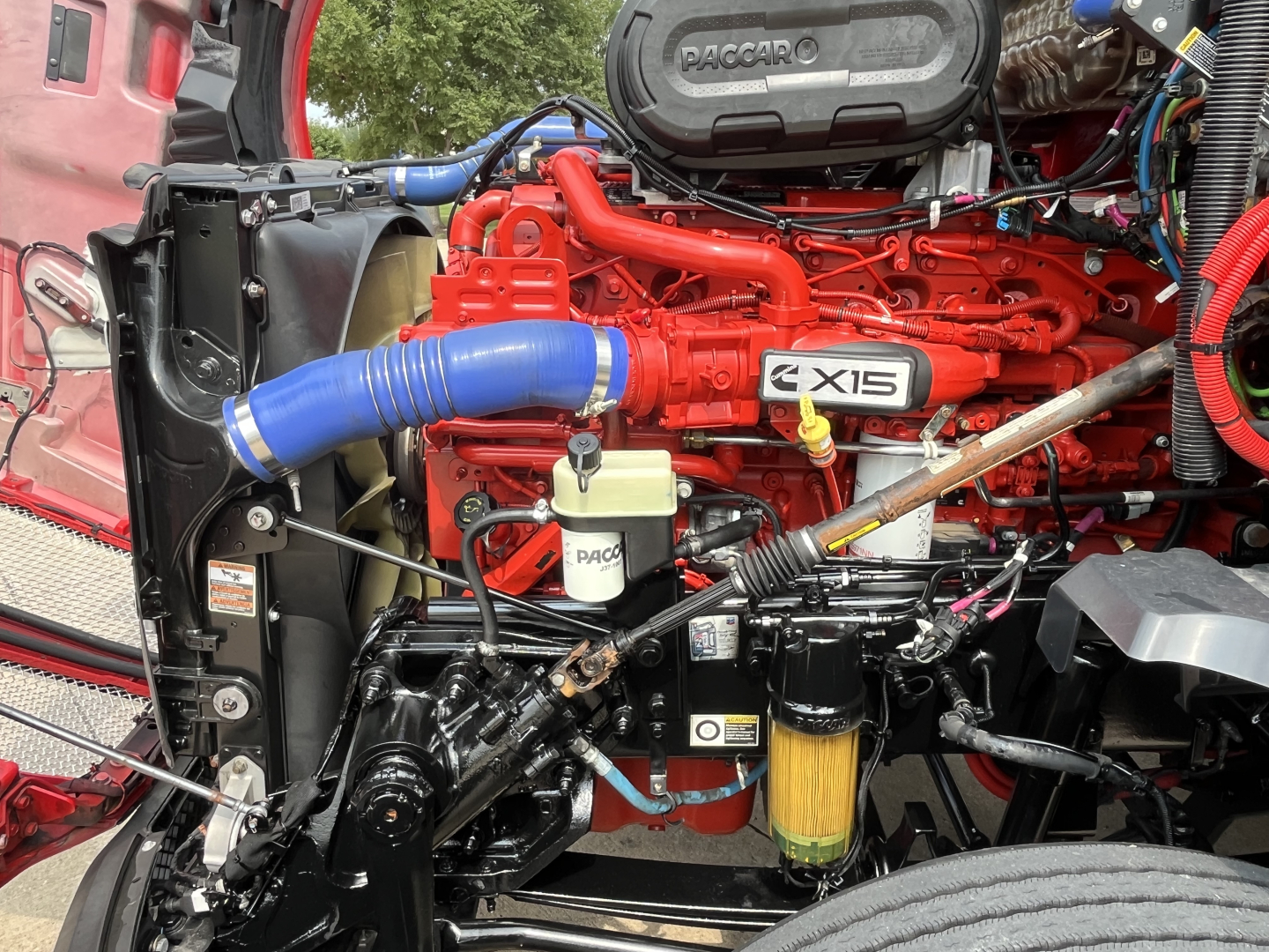 Kenworth T680 engine bay with red block and blue hose, showing RACCARO air intake, X15 badge, white PASCO/PACCAR reservoir, yellow oil filter, and various hoses and wiring.