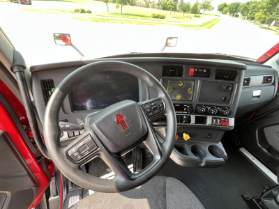 Cab interior of a 2024 Kenworth T680 with steering wheel, dashboard, and center console visible; features include a large digital display, various control panels, cup holders, and a red door frame.