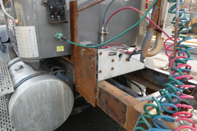Truck chassis area showing metal frame and air lines; battery box and wiring visible on a rusty frame rail connected to a large cylindrical tank or air reservoir.