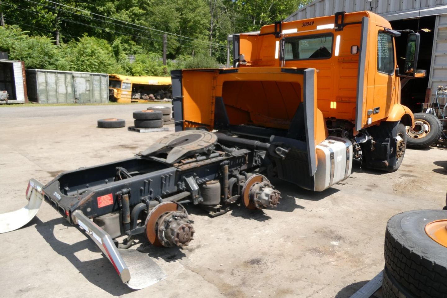 Wheel-equipped Volvo VNR64T300 daycab chassis with cab removed, orange cab and engine bay visible, exposed drivetrain and rear axles, daytime outdoor lot