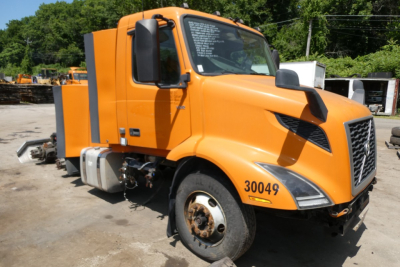 Orange 2019 Volvo VNR64T300 conventional daycab truck showing front-left view with missing front wheel; cab window stickers visible on windshield.