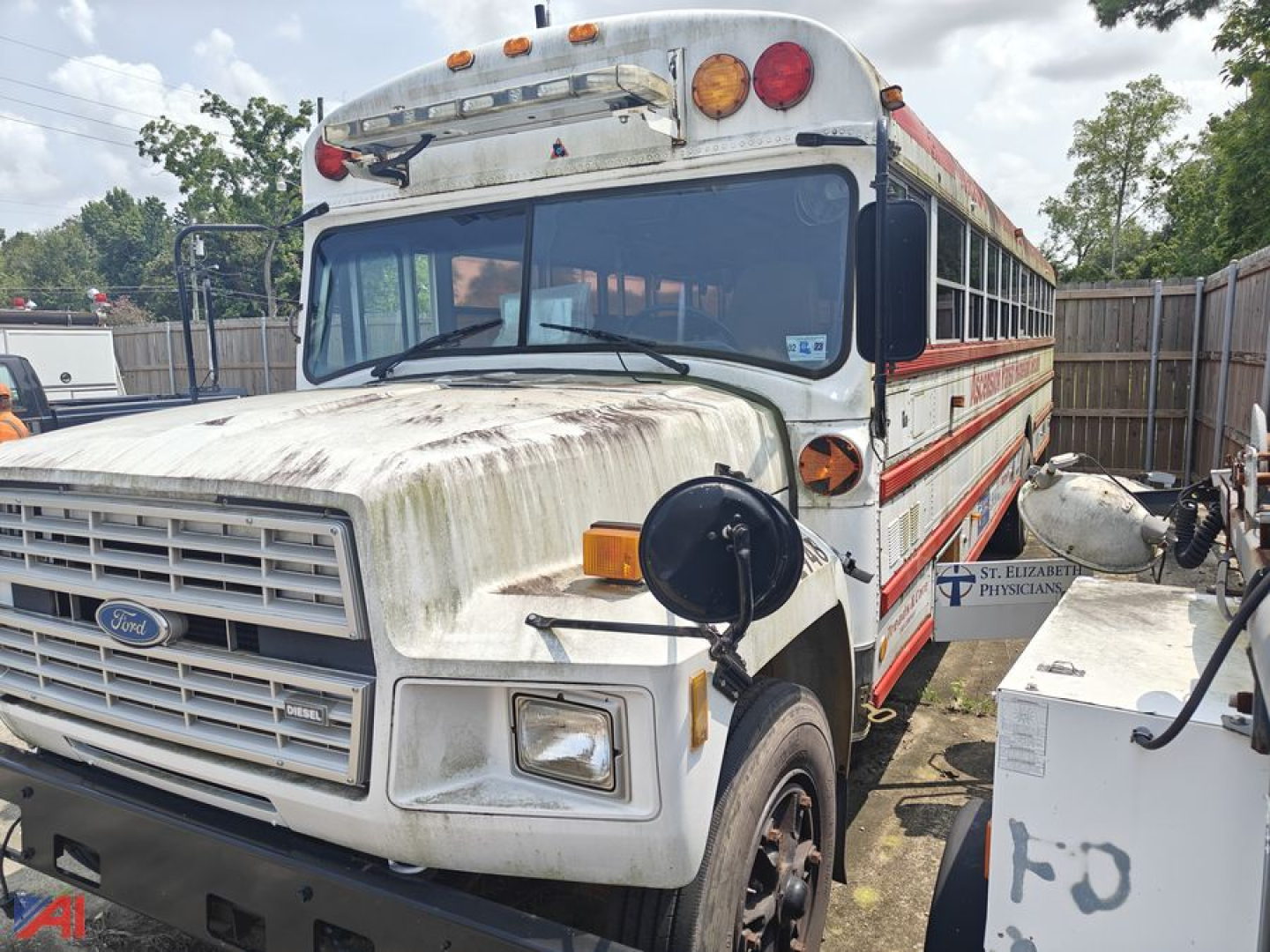 Ford bus nose and side view showing weathered white body with red stripe; Ford emblem on grille, round amber turn signals, front bumper, school bus windows, and passengers door. Text not readable due to angle.
