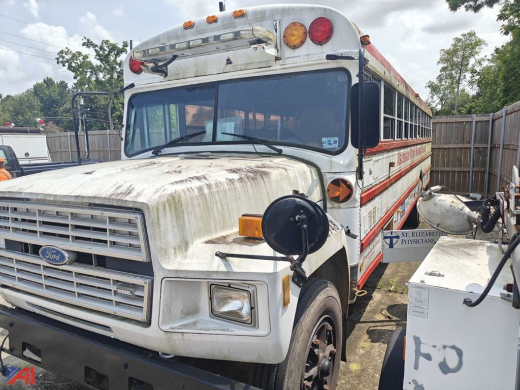 Ford bus nose and side view showing weathered white body with red stripe; Ford emblem on grille, round amber turn signals, front bumper, school bus windows, and passengers door. Text not readable due to angle.