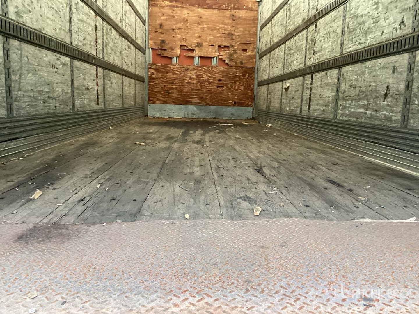 Trailer interior showing empty cargo area with wooden bulkhead, metal walls, and weathered floor; rear open with diamond plate threshold.