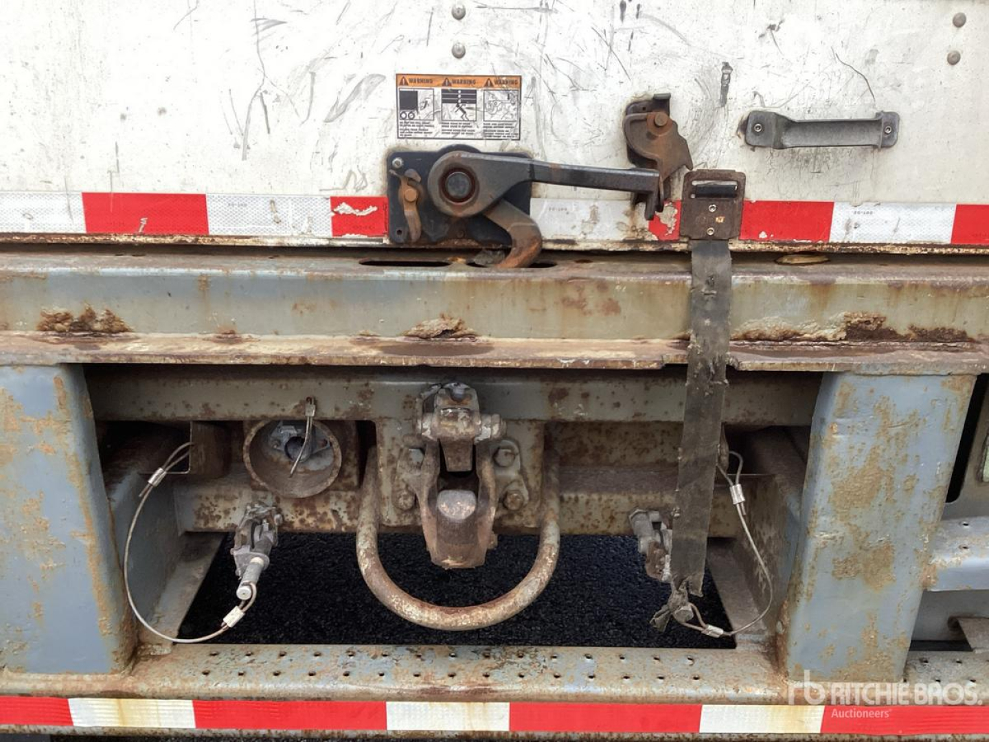 Trailer hitch and frame of a used van trailer showing a circular hitch ring, two side locking mechanisms, and rusted metal with red/white reflective tape.