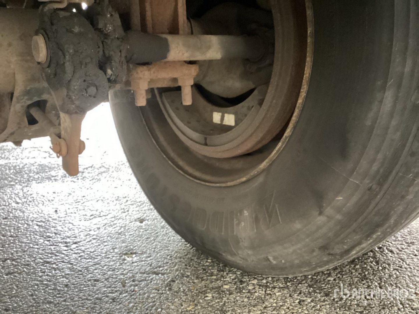 Trailer wheel and suspension assembly with tire, axle, and leaf spring visible; U-bolt, brake components, and mud residue on wheel area.