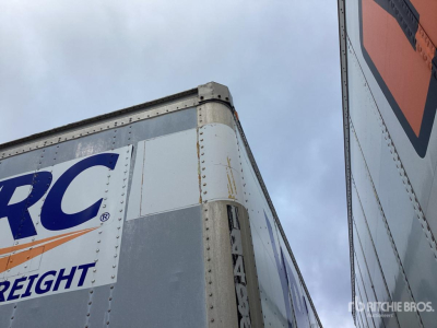 Truck trailer corner showing RC_? branding on a white-and-blue panel with riveted metal edge and orange safety stripe.