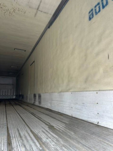 Inside view of a refrigerated trailer, metal floor with rails, insulated sidewall with partial blue text and a door at far end.